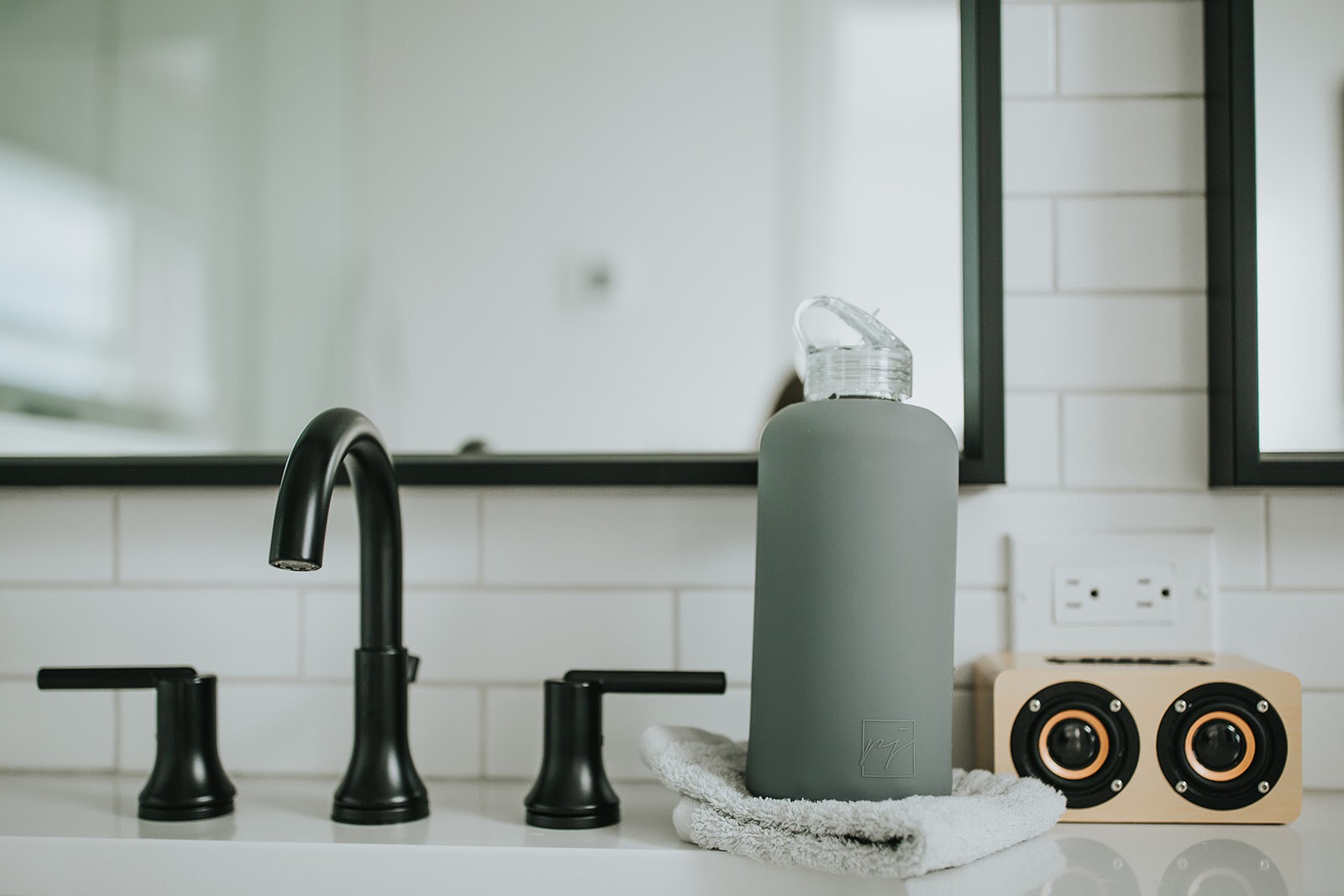 One litre glass water bottle with grey protective silicone sleeve and clear folding straw lid. Lifestyle picture of grey water bottle sitting on folded towel next to a black faucet with white subway tiles and black framed mirror - lifewithPandJ