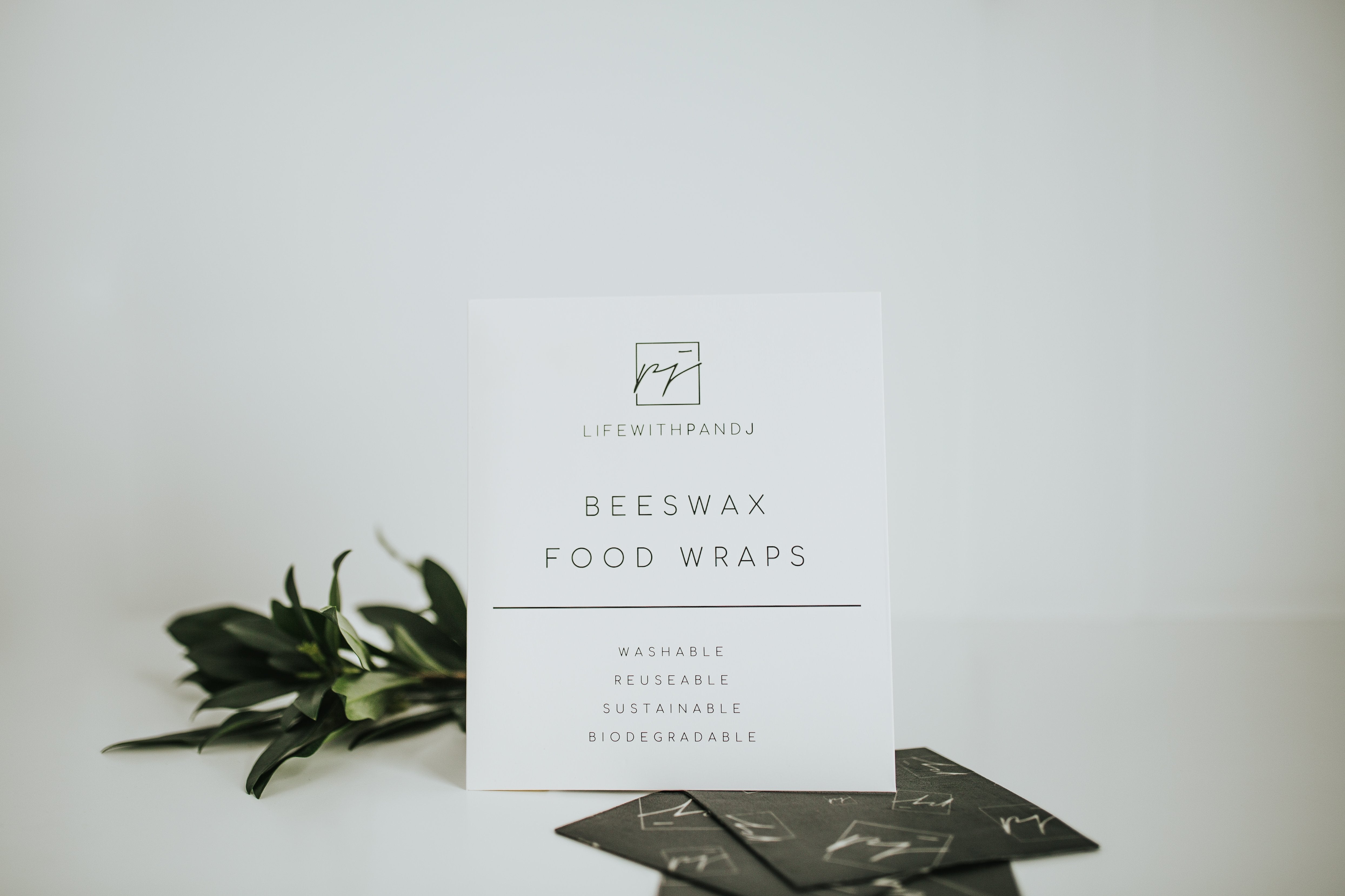 Beeswax Food Wraps with sleek and minimalist white packaging that reads, "washable, reusabe, sustainable, and biodegradable". 3 black food wraps are laying flat on a white background with greenery - lifewithPandJ