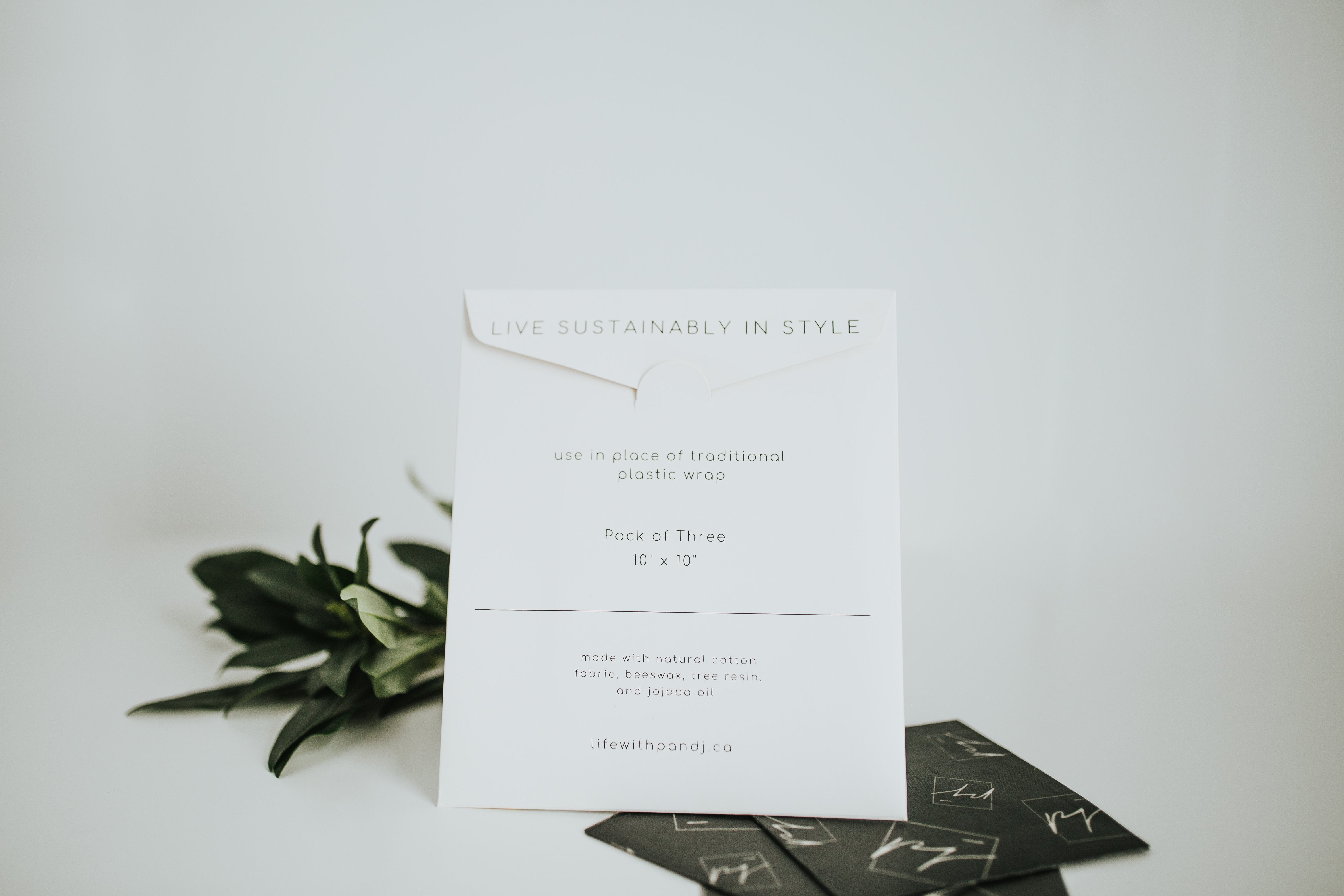 Beeswax Food Wraps with sleek and minimalist white packaging that reads, "pack of 3 10"x10", list of natural ingredients and, live sustainably in style". 3 black food wraps are laying flat on a white background with greenery - lifewithPandJ