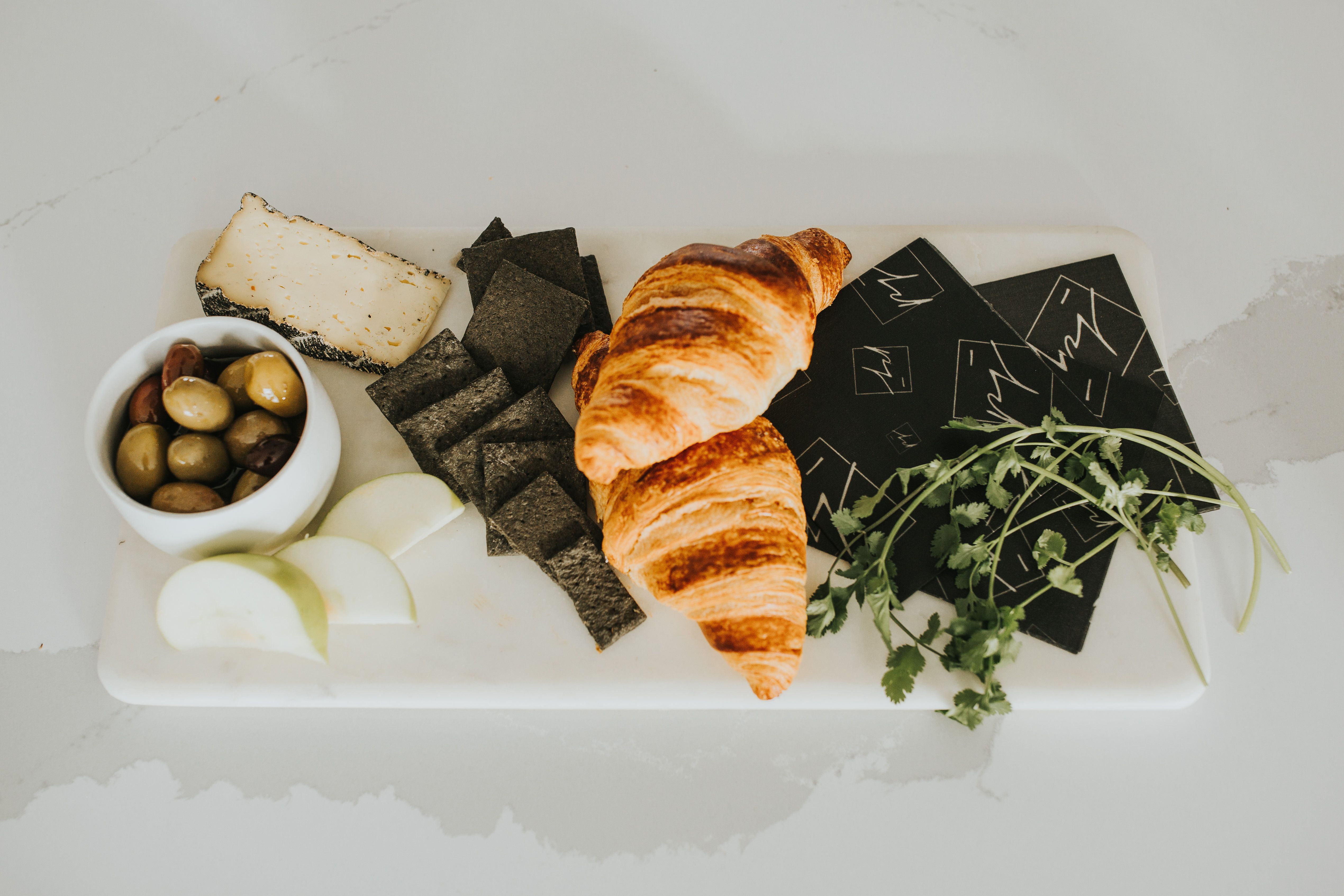 A beatiful charcuterie board made up of cheese, olives, black crackers, green apple, croissants and cilantro. 3 black beeswax wraps are laying on a marble countertop - lifewithPandJ