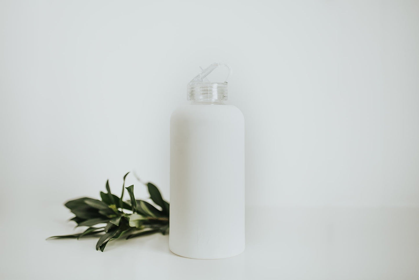 1L glass water bottle with white protective silicone sleeve and clear folding straw lid with a white background and greenery - lifewithPandJ