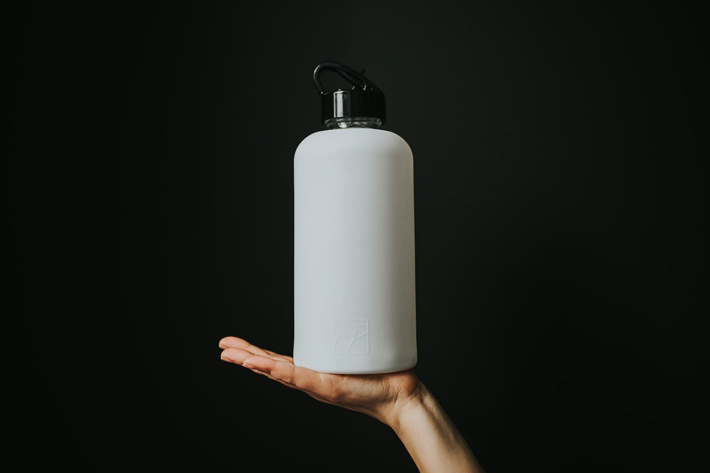 1L glass water bottle with white protective silicone sleeve and high gloss black lid on a black background and being held by a woman's hand - lifewithPandJ