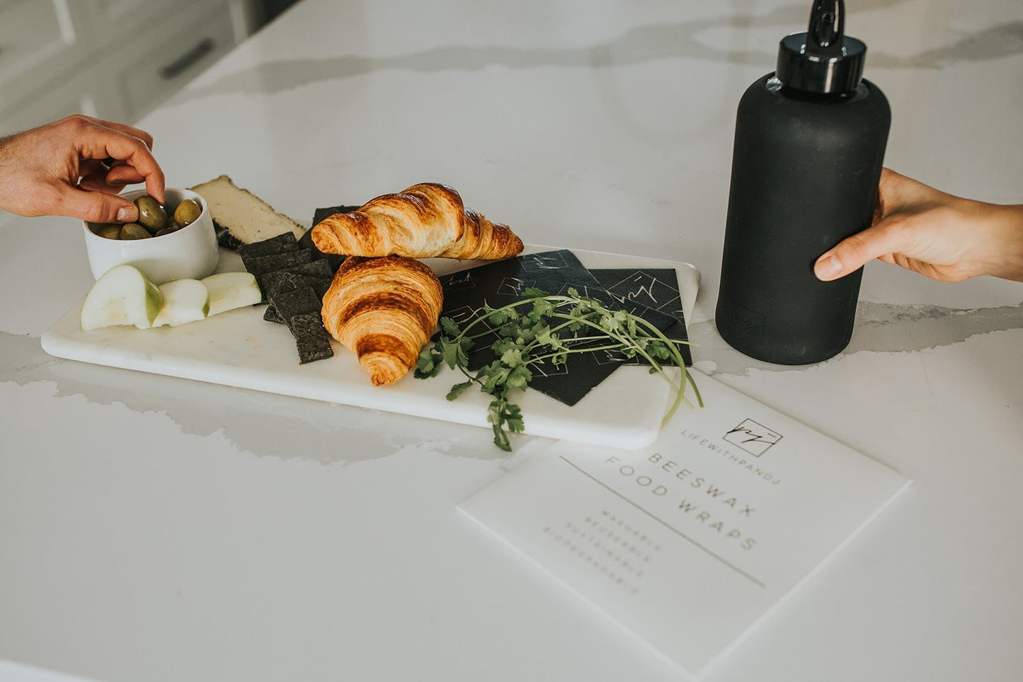 1L glass water bottle with high gloss black folding straw lid beside a beautiful charcuterie board made up of cheese, olives, black crackers, green apple, croissants and cilantro. 3 black beeswax wraps are laying on a marble countertop with the minimalist white packaging of beeswax food wraps - lifewithPandJ