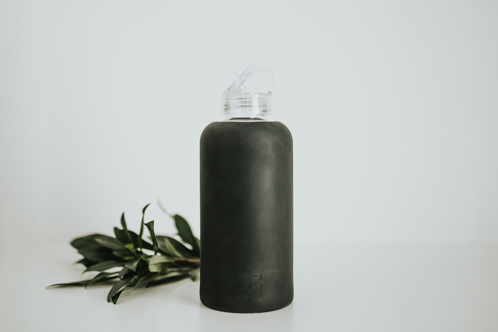 1L glass water bottle with Onyx protective silicone sleeve and clear folding straw lid on a white background with greenery - lifewithPandJ