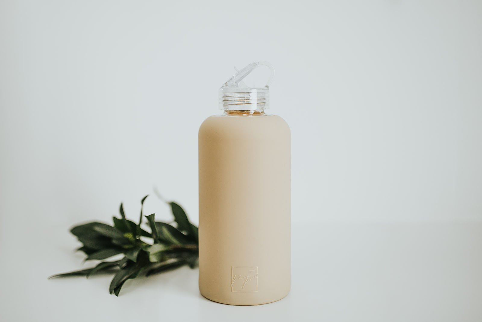 One litre glass water bottle with nude, sand coloured protective silicone sleeve and clear folding straw lid. White background with green foliage behind water bottle - lifewithPandJ
