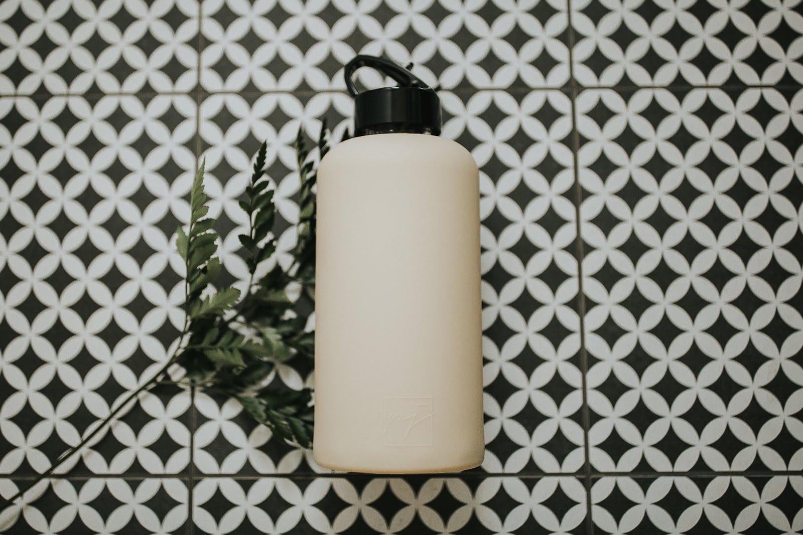 One litre glass water bottle with nude, sand coloured protective silicone sleeve and high gloss black folding straw lid. On a black and white tiled background with green foliage - lifewithPandJ