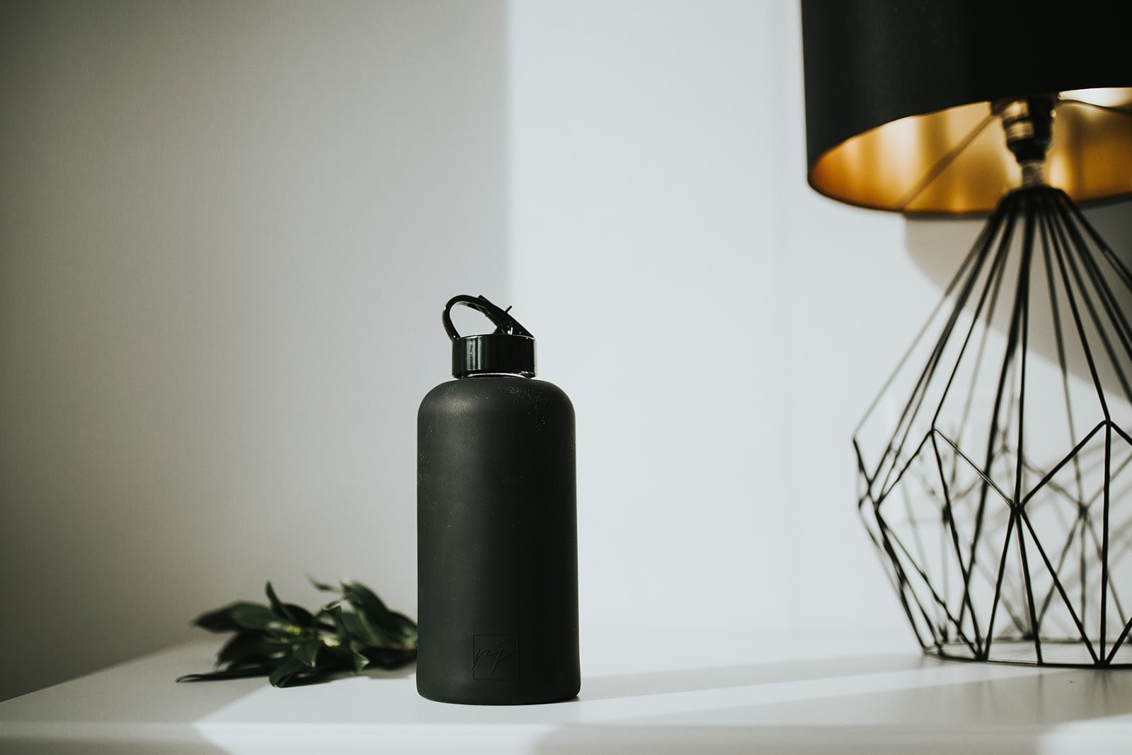 1L glass water bottle with black Onyx protective silicone sleeve and high gloss black folding straw lid on a bedside night stand with a black and gold designer lamp shade and greenery- lifewithPandJ