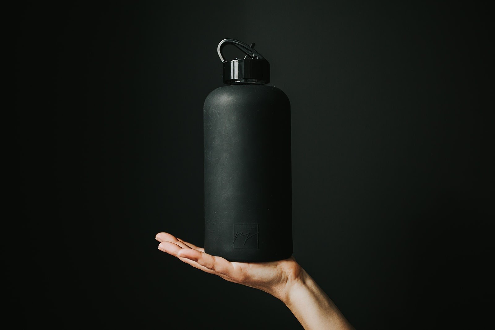 1L glass water bottle with black Onyx protective silicone sleeve and high gloss black folding straw lid being held up by a woman's hand on a black background - lifewithPandJ