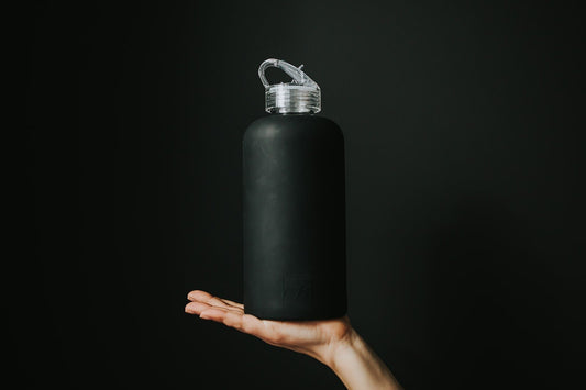 1L glass water bottle with black Onyx protective silicone sleeve and clear folding straw lid being held up by a woman's hand on a black background - lifewithPandJ 