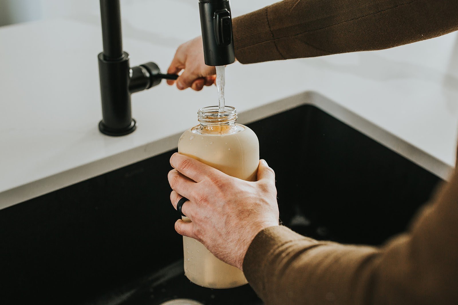 One litre glass water bottle with nude, sand protective silicone sleeve. Lifestyle photography of man holding bottle and filling up from black faucet - lifewithPandJ