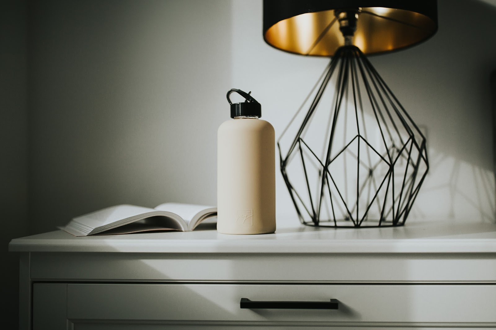 One litre glass water bottle with nude, sand coloured protective silicone sleeve and high gloss black folding straw lid. Lifestyle photography, bottle on top of white dresser with open book and black and gold geometric lamp - lifewithPandJ