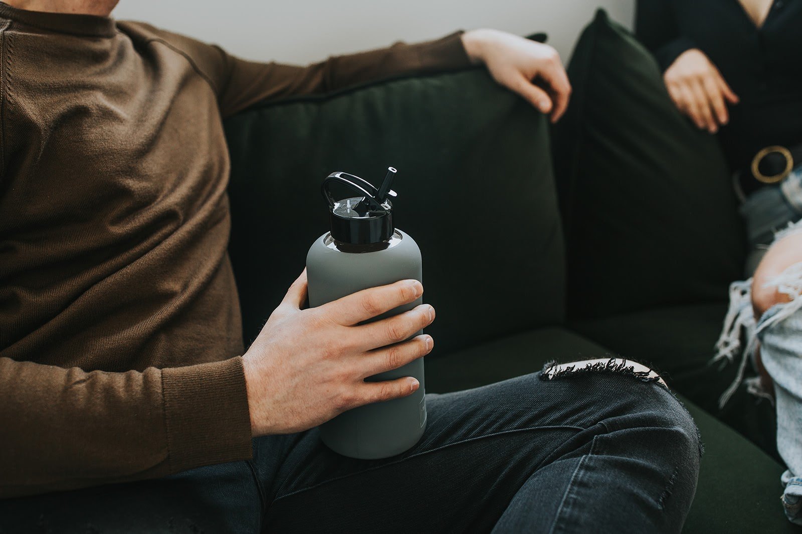 One litre glass water bottle with grey protective silicone sleeve and black folding straw lid. Lifestyle photography of two people sitting on dark green couch, one is holding grey water bottle with high gloss folding black lid- lifewithPandJ